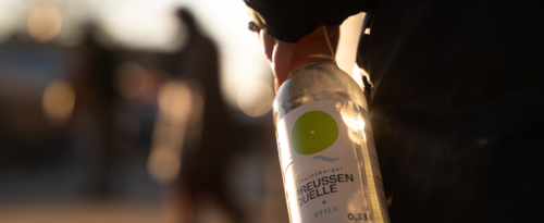 jemand trägt lässig in einer Hand neben dem Körper eine Glasflasche im Gegenlicht mit dem Etikett der Rheinsberger Preussenquelle