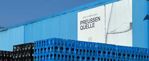 Die Produktionshalle in hellblau mit einem weißen Aufdruck und dem Logo nebst einer Falsche Rheinsberger Preussenquelle vor einem blauen Himmel mit davor getürmten Kästen der Marke