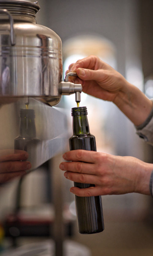 zwei Hände eine Ölflasche haltend und Öl aus einem Edelstahlbehälter über Hahn abfüllend
